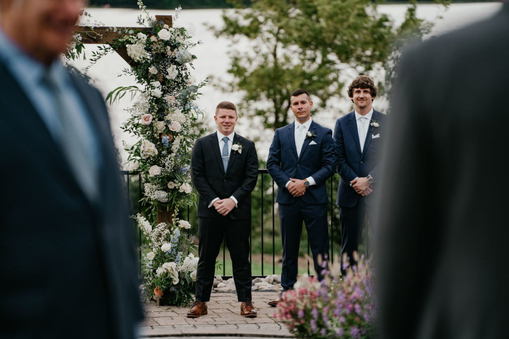 groom watching his bride walk down the aisle framed by a floral arch