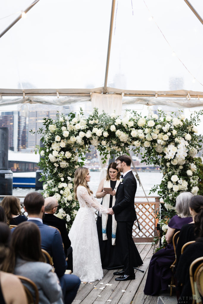 Bride and Groom under white and green floral arch filled with hydrangea, roses and orchids